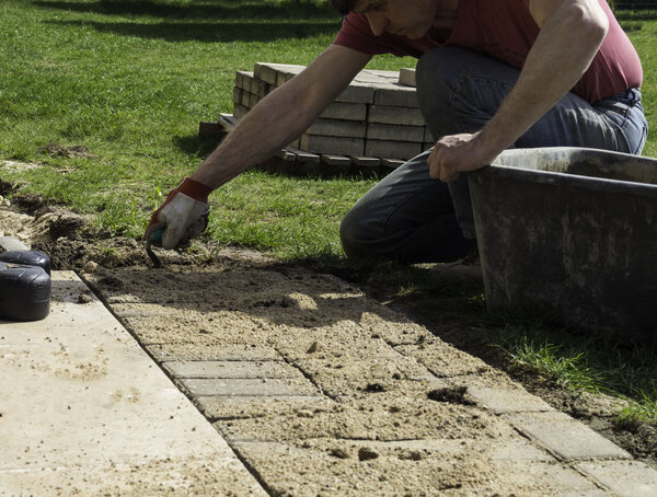Handwerker verlegt Gehwegplatten auf Sanduntergrund im Garten.